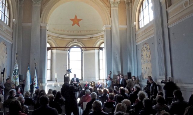 Pohled na průčelí kostela s mluvčím vepředu a publikem sedícím na židlích